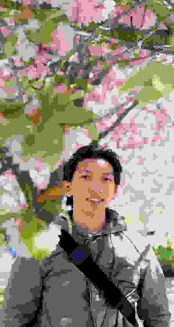 A young man stands smiling in front of blossoming cherry trees with pink flowers.