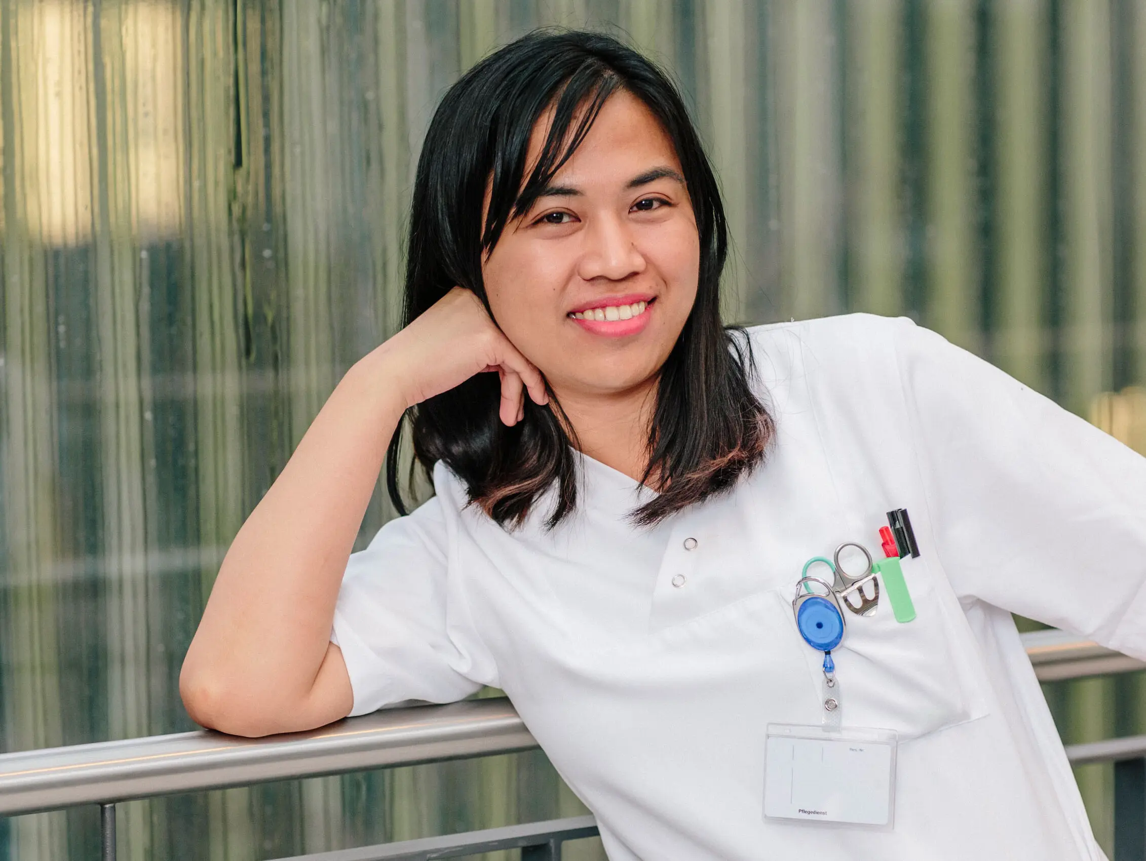 A smiling person in a white doctor's coat leans relaxed against a railing in front of a modern glass wall.
