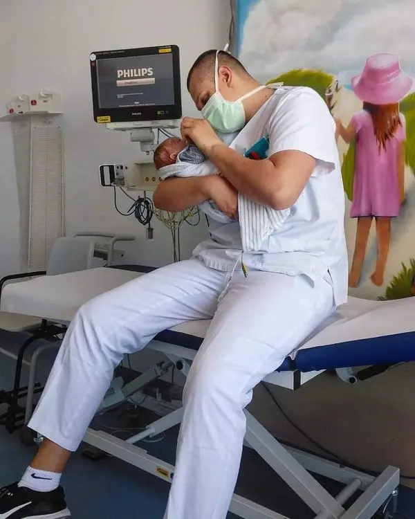 A nurse in the medical room holds a newborn baby in his arms and looks at it. A screen and a mural can be seen in the background.