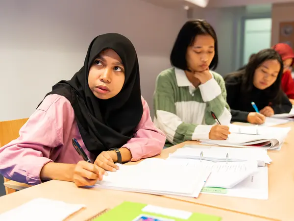 A group of young women sit at a table, focused on their notes and writing as they take part in a class.