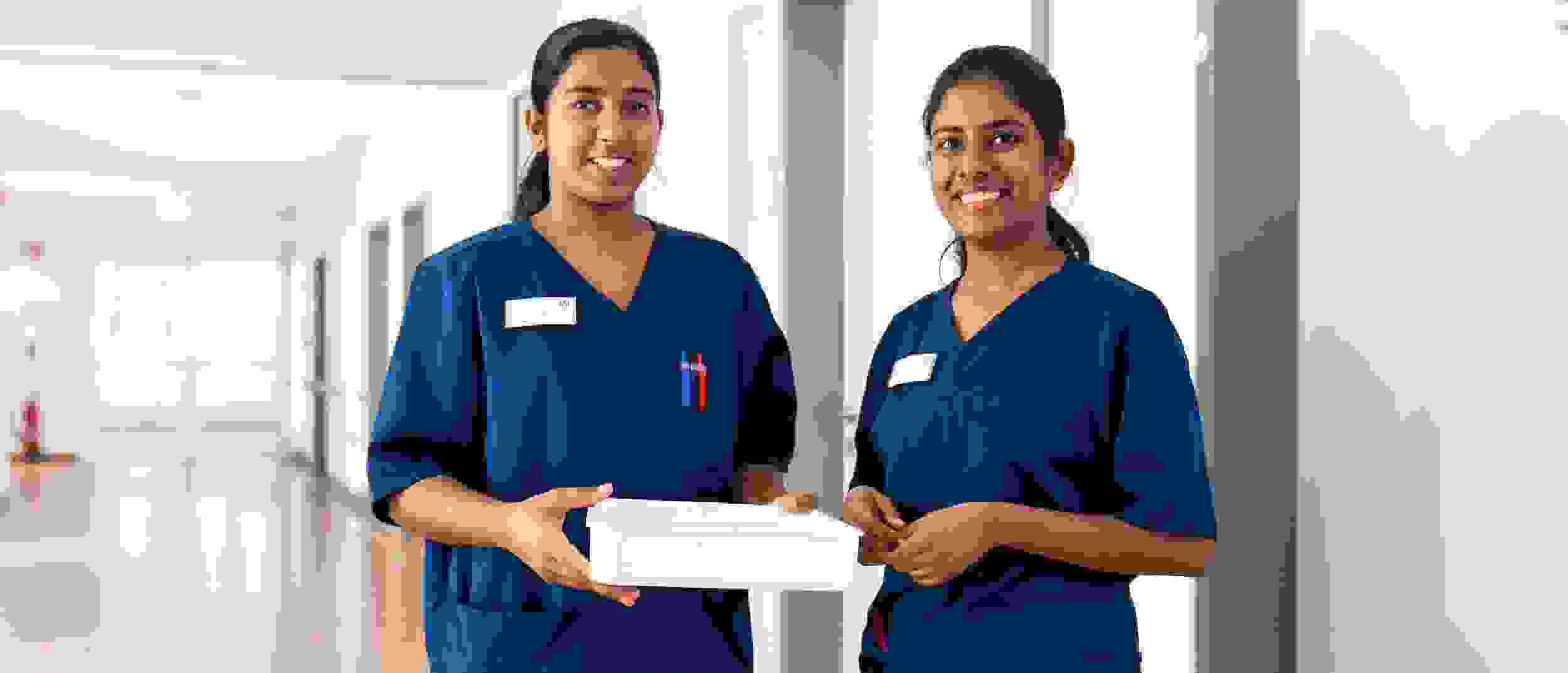Two nurses in blue uniforms stand in the hospital corridor holding a white box.