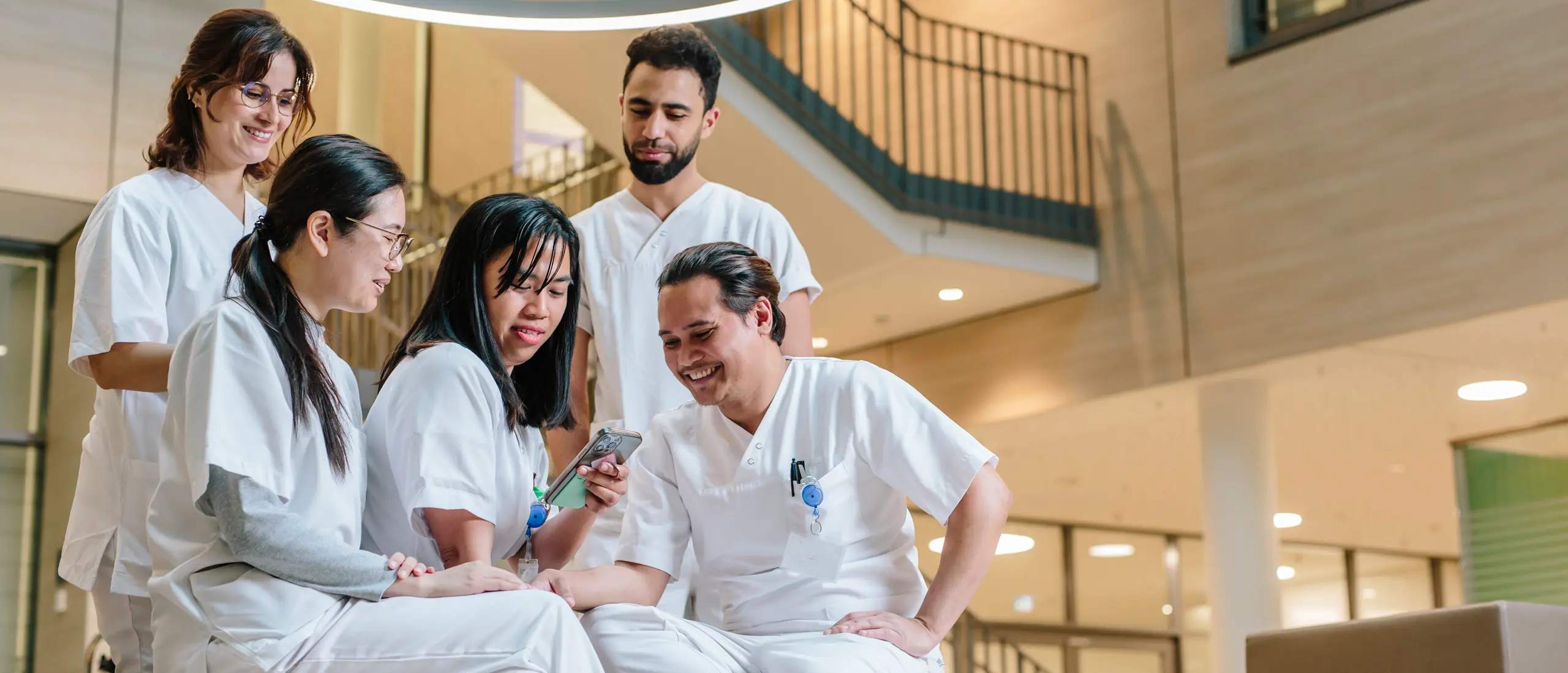 Cinq professionnels de la santé en blouse blanche assis ensemble sur un canapé et regardant un téléphone portable dans un intérieur moderne.