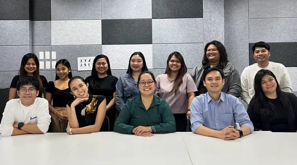 A group of 13 people sit and stand in front of a gray wall in a modern meeting room. They smile and pose together.