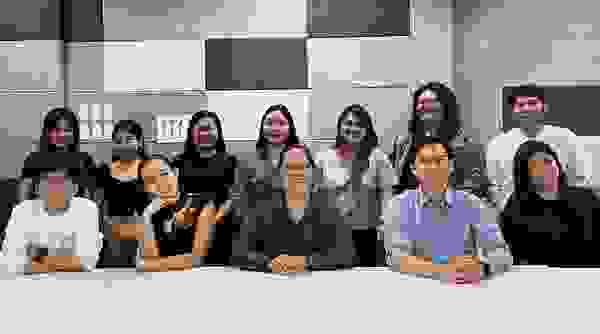 A group of 13 people sit and stand in front of a gray wall in a modern meeting room. They smile and pose together.