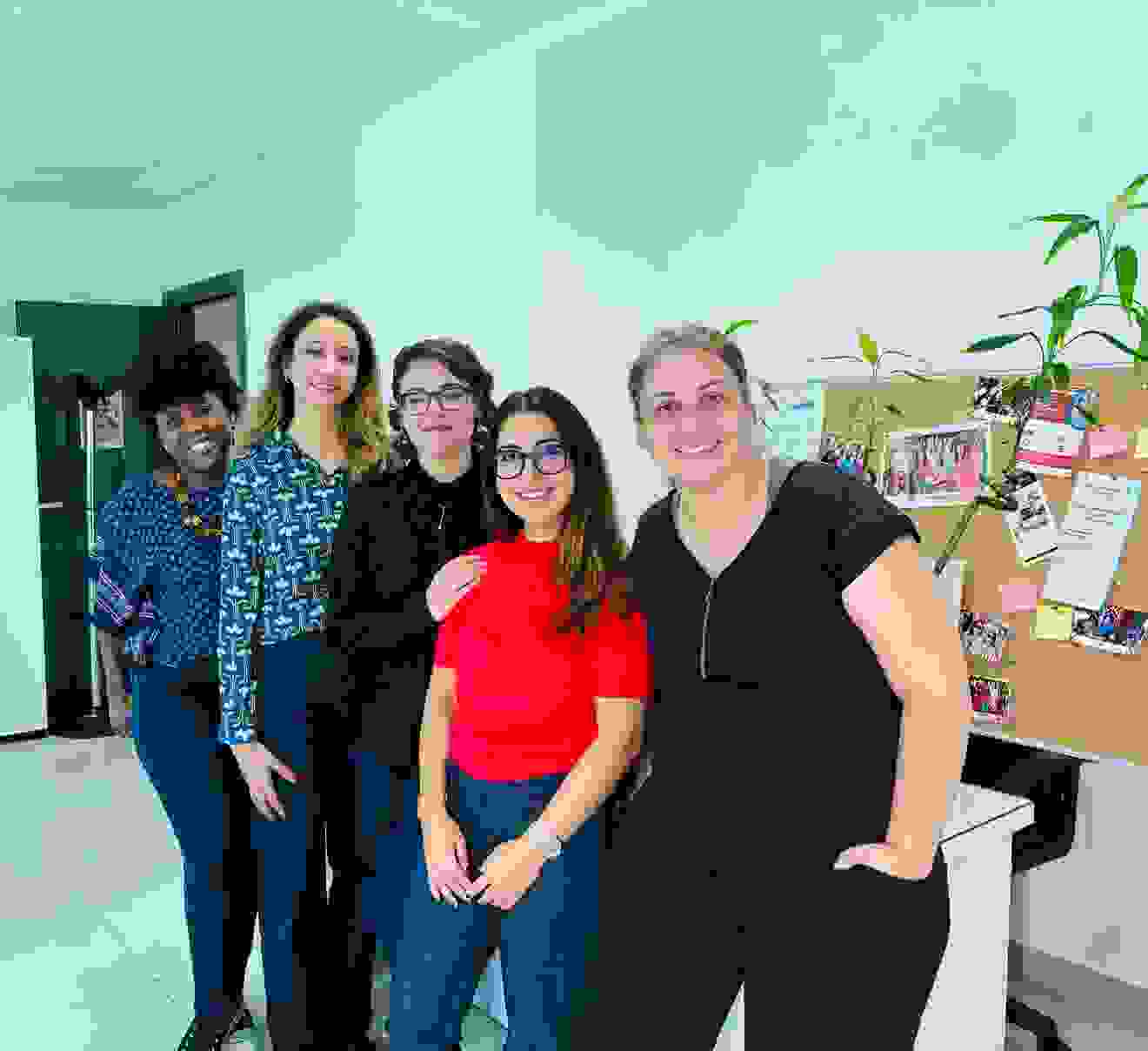 Cinq femmes se tiennent dans un bureau, souriant et posant ensemble. Derrière elles, un tableau mural avec des notes et des plantes.