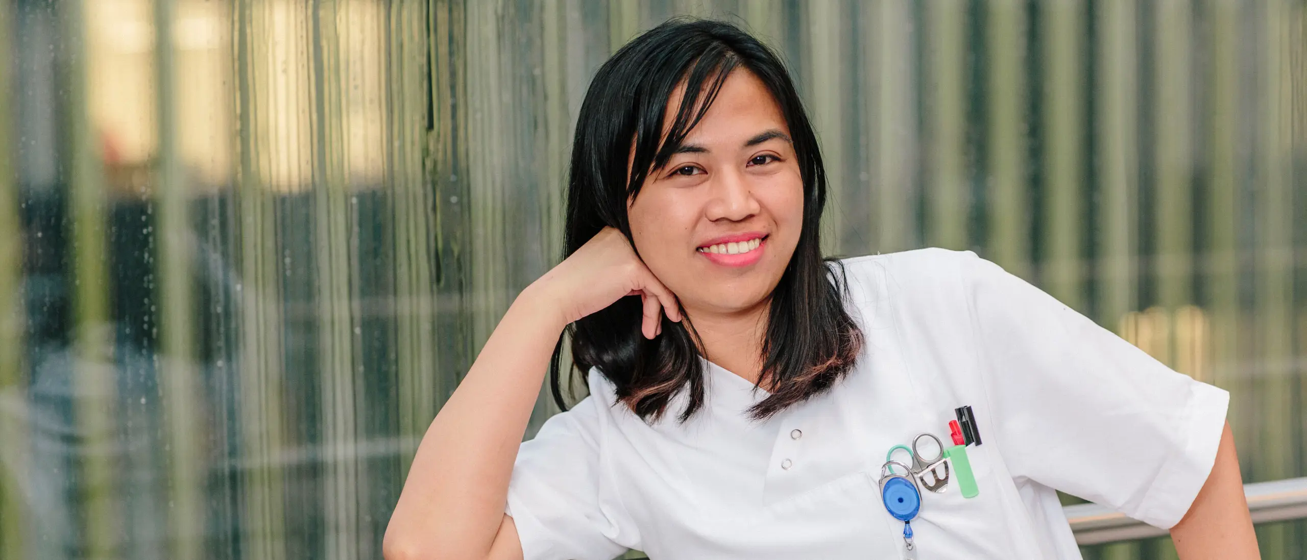 A smiling person in a white doctor's coat leans relaxed against a railing in front of a modern glass wall.