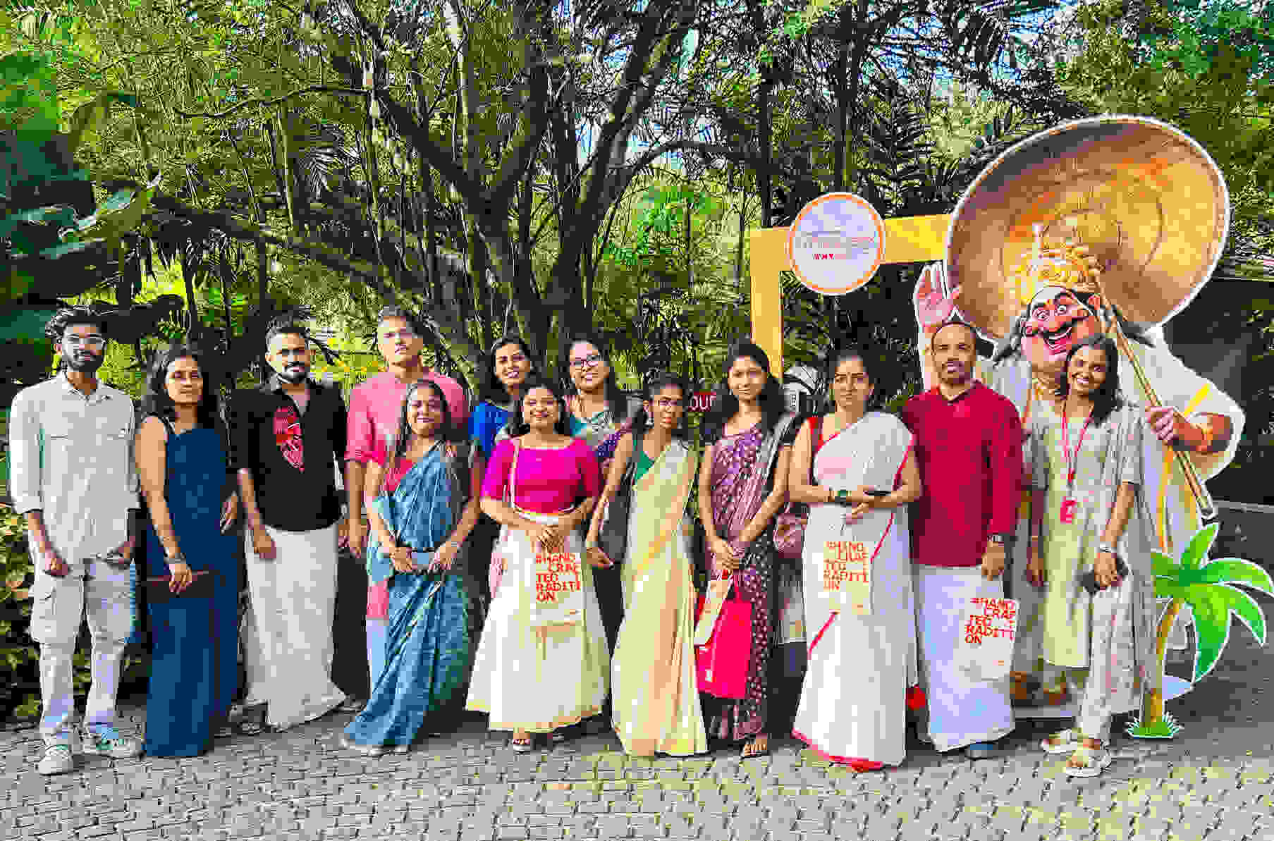 A group of people in traditional Indian dress pose in front of a green background and a colorful, carved character.