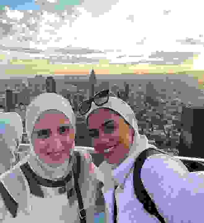 Two women with headscarves pose for a selfie with a city view in the background at sunset.