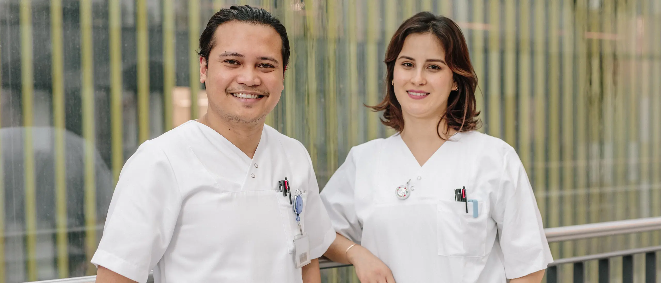 Deux professionnels de la sant&eacute; souriants, en blouse blanche, se tiennent devant un mur de verre moderne &agrave; rayures verticales.