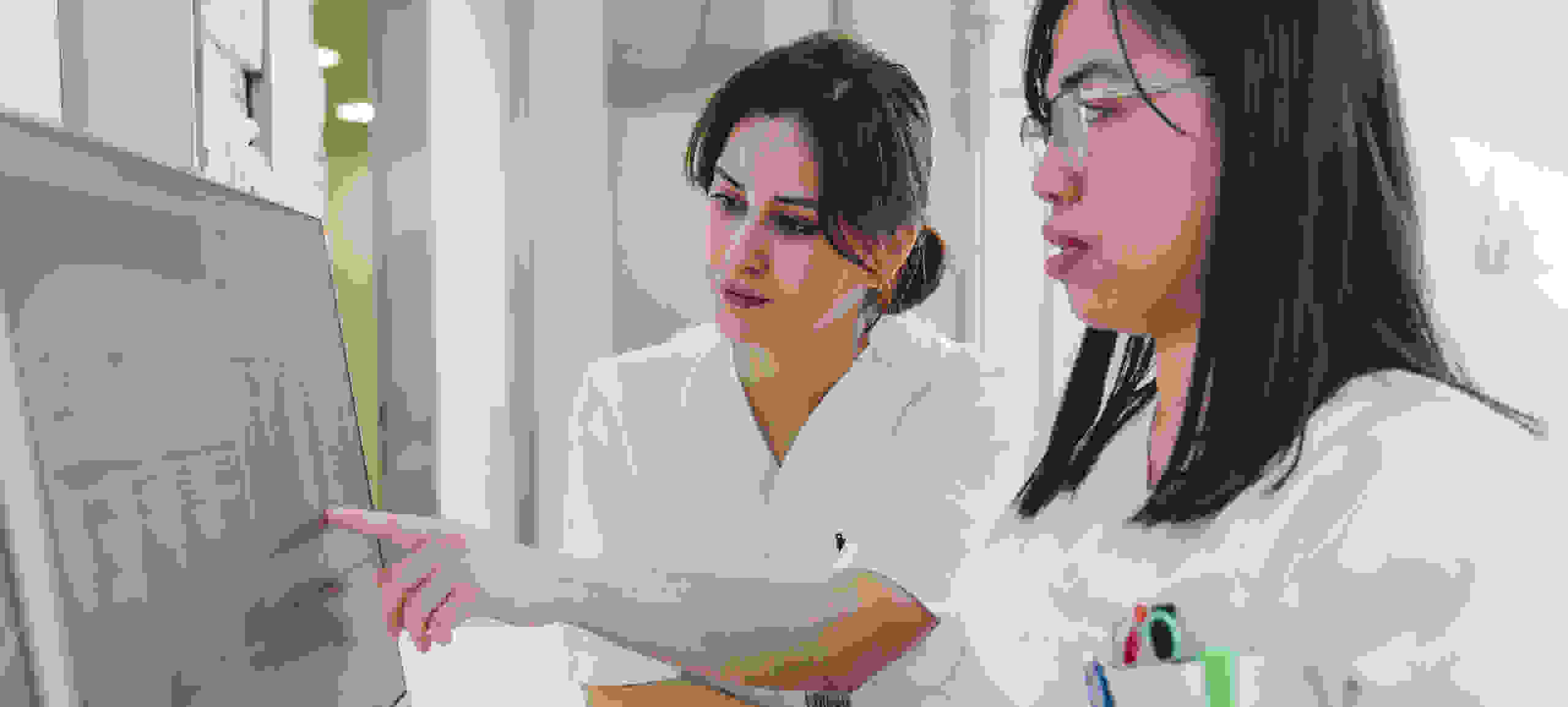 Deux femmes vêtues de vêtements de laboratoire blancs regardent ensemble un écran d'ordinateur et le montrent du doigt.