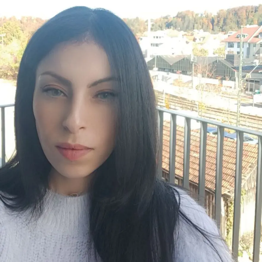 Gros plan d'un visage aux cheveux longs et noirs, portant un pull de couleur claire. Des b&acirc;timents et des arbres sont visibles &agrave; l'arri&egrave;re-plan.