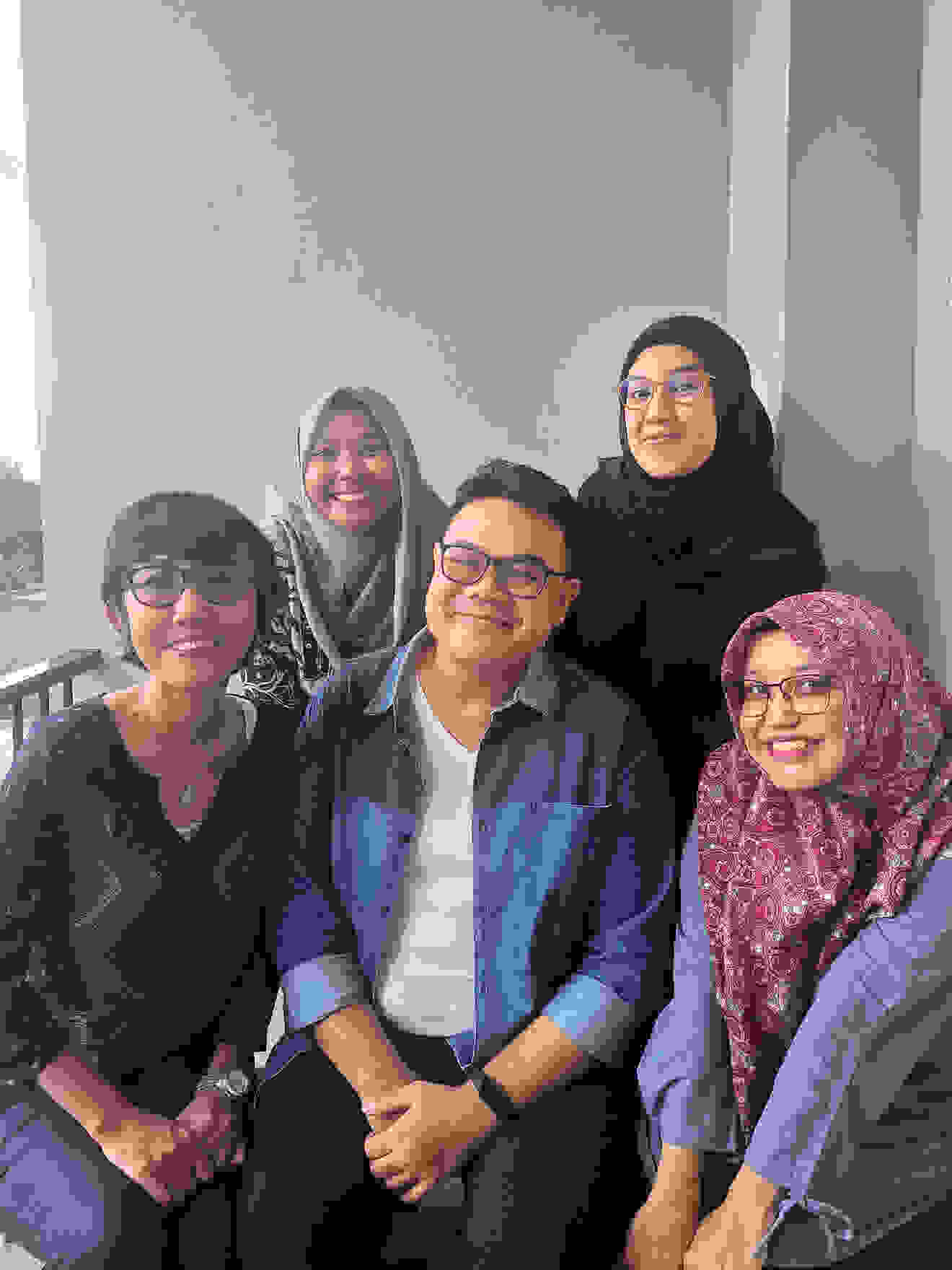 Five people pose smiling in front of a wall, two are sitting and three are standing behind them.