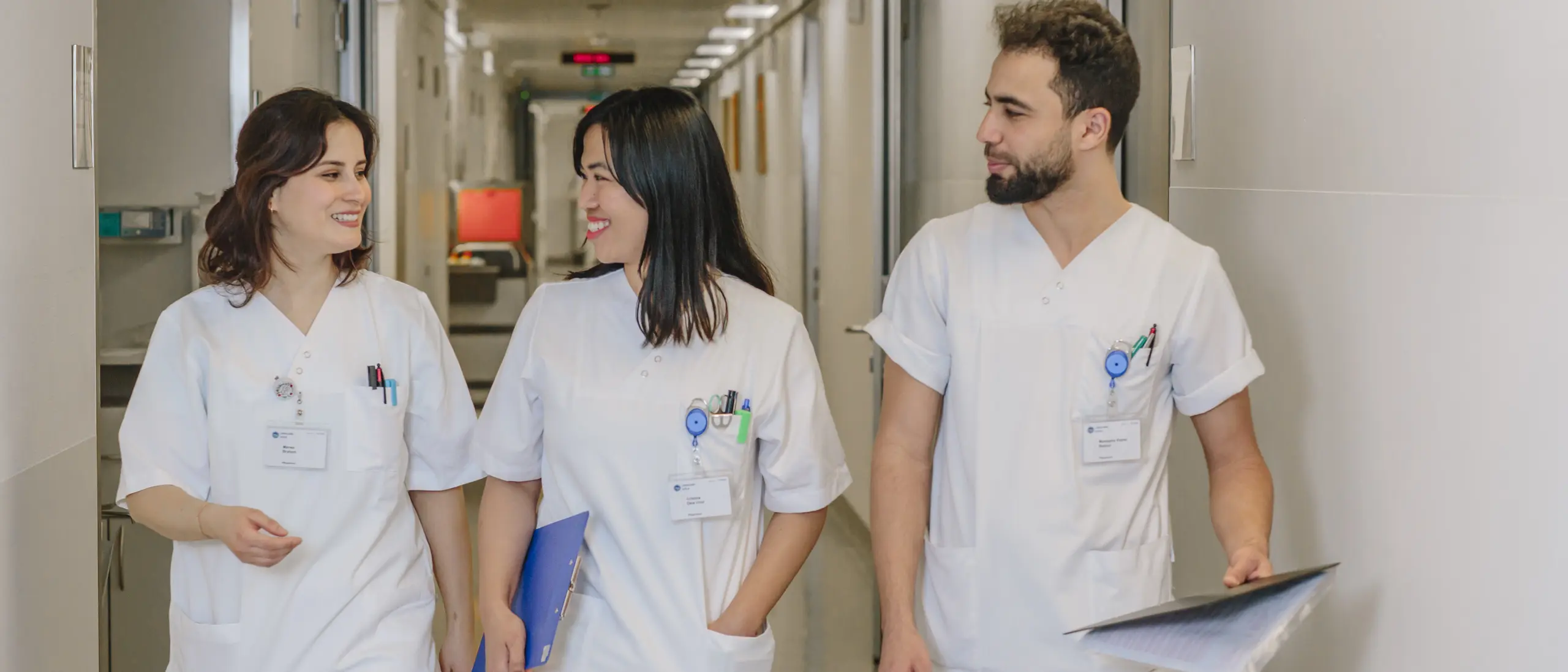 Trois professionnels de la santé en uniforme blanc marchent dans un couloir d'hôpital, conversant et souriant.