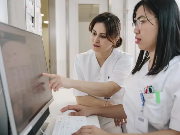 Deux femmes vêtues de vêtements de laboratoire blancs regardent ensemble un écran d'ordinateur et le montrent du doigt.