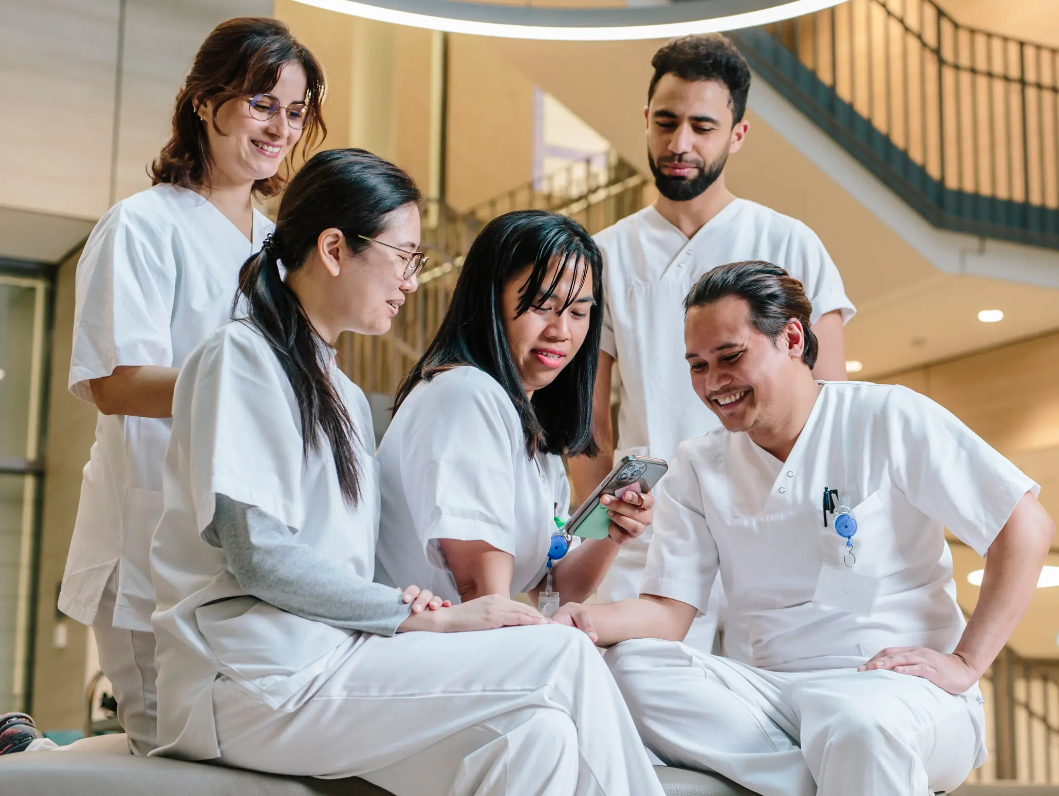 Cinq professionnels de la santé en blouse blanche assis ensemble sur un canapé et regardant un téléphone portable dans un intérieur moderne.