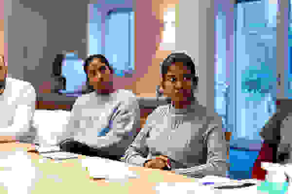 Deux femmes sont assises à une table dans une salle de réunion. L'une porte un pull bleu clair structuré, l'autre un pull bleu à col roulé, et toutes deux écoutent attentivement. Un homme est également visible sur l'image, mais son regard n'est pas dirigé vers l'appareil photo.