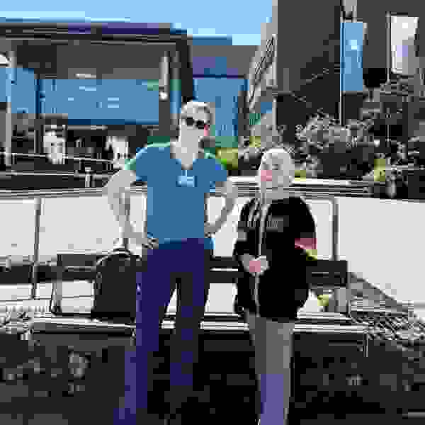 Deux personnes se tiennent devant un bâtiment moderne. L'une porte une chemise bleue et des lunettes de soleil, l'autre un sweat-shirt noir à rayures colorées. Toutes deux sourient légèrement.