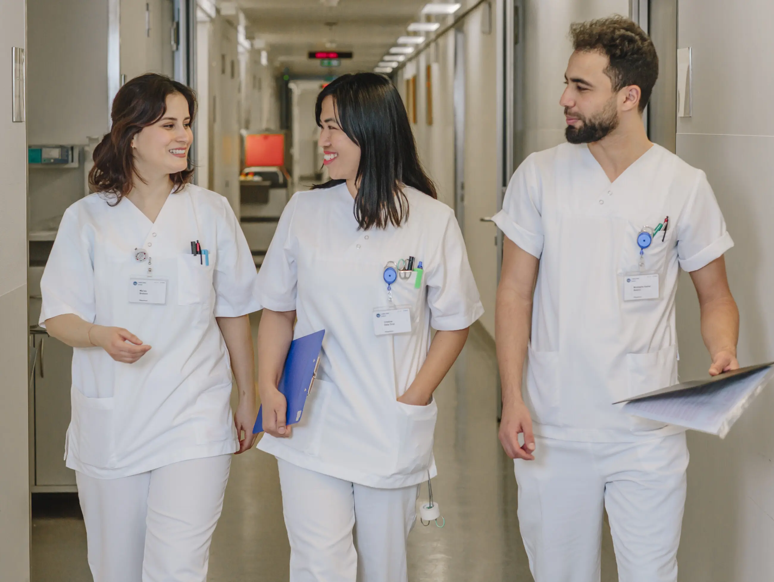 Trois professionnels de la santé en uniforme blanc marchent dans un couloir d'hôpital, conversant et souriant.