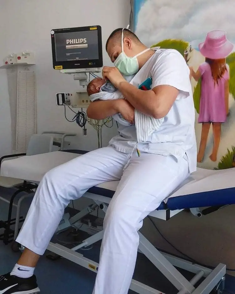 Seorang perawat di ruang medis menggendong bayi yang baru lahir dan melihatnya. Sebuah layar dan mural terlihat di latar belakang.