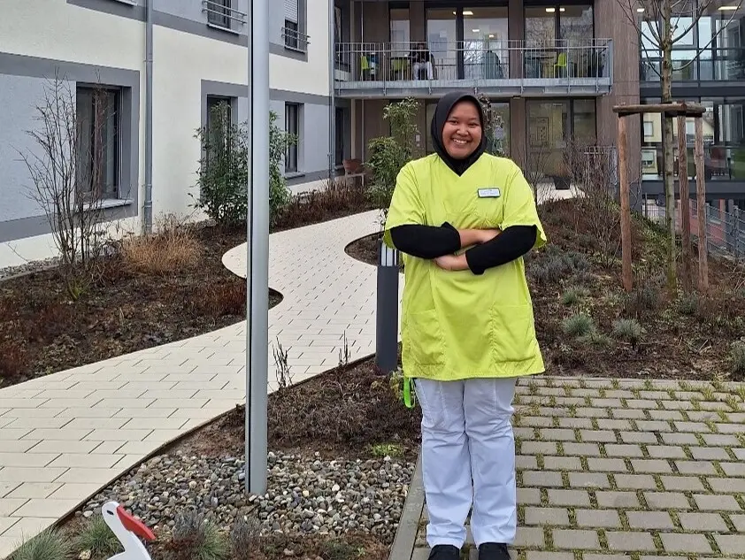 Seseorang dengan pakaian kerja berwarna kuning berdiri di halaman dalam sebuah bangunan modern, dikelilingi oleh tanaman dan trotoar. Jendela dengan balkon dan tiang bendera terlihat di latar belakang.