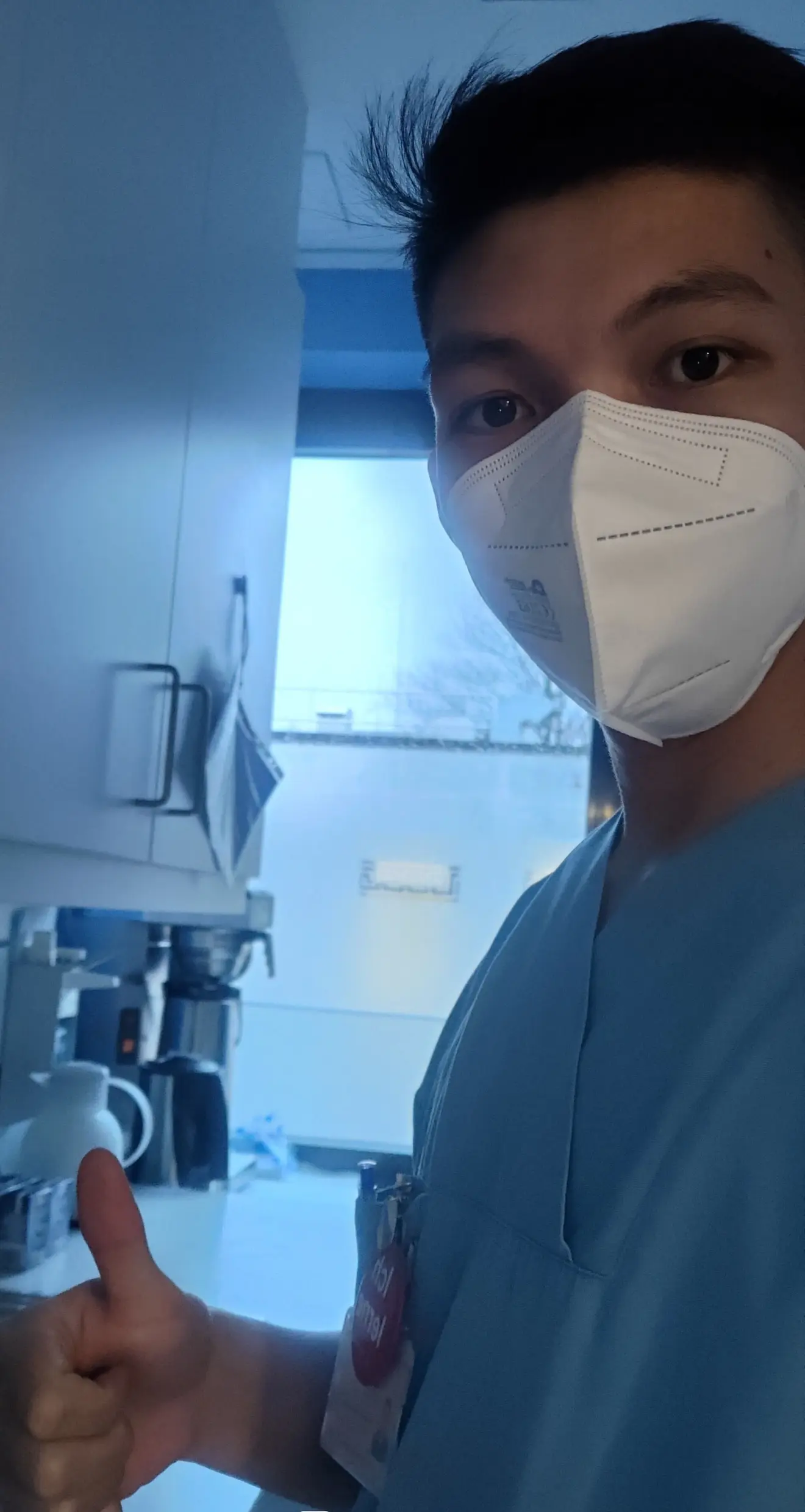 Seorang pria muda dengan pakaian kerja berwarna biru dan masker medis, mengacungkan jempol, berdiri di lingkungan klinik yang cerah.