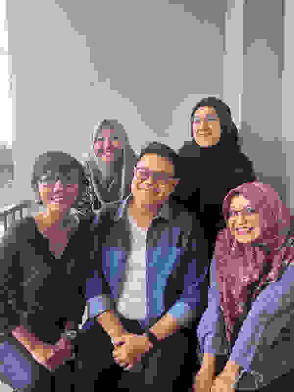 Lima orang berpose tersenyum di depan dinding, dua orang duduk dan tiga orang berdiri di belakang mereka.