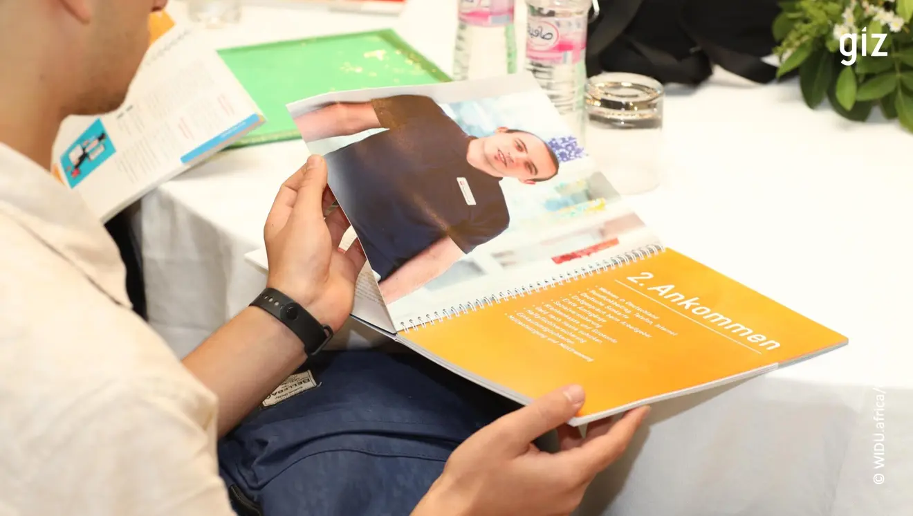 A person is holding an open booklet with a picture of a man in a dark shirt. The title on one page reads “2nd arrival” in German. Water bottles and leaves can be seen in the background.