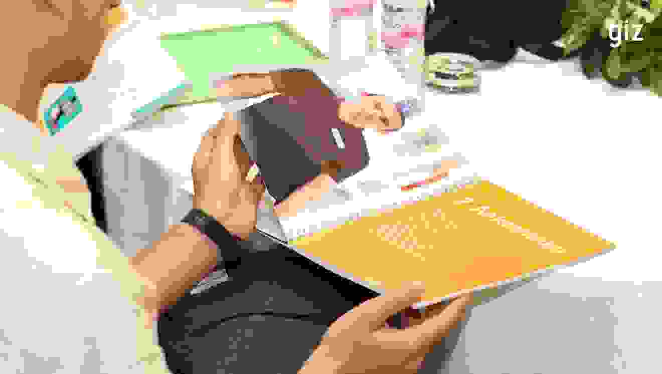 A person is holding an open booklet with a picture of a man in a dark shirt. The title on one page reads “2nd arrival” in German. Water bottles and leaves can be seen in the background.