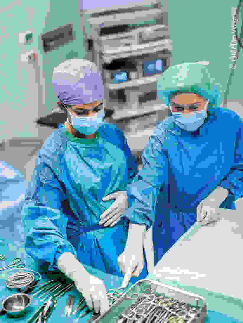 Dua ahli bedah dengan gaun dan masker bedah berwarna biru berdiri di ruang operasi dan menyiapkan instrumen bedah.