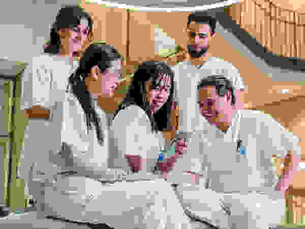 Lima orang profesional medis berjas putih duduk bersama di sofa sambil melihat ponsel dalam interior yang modern.