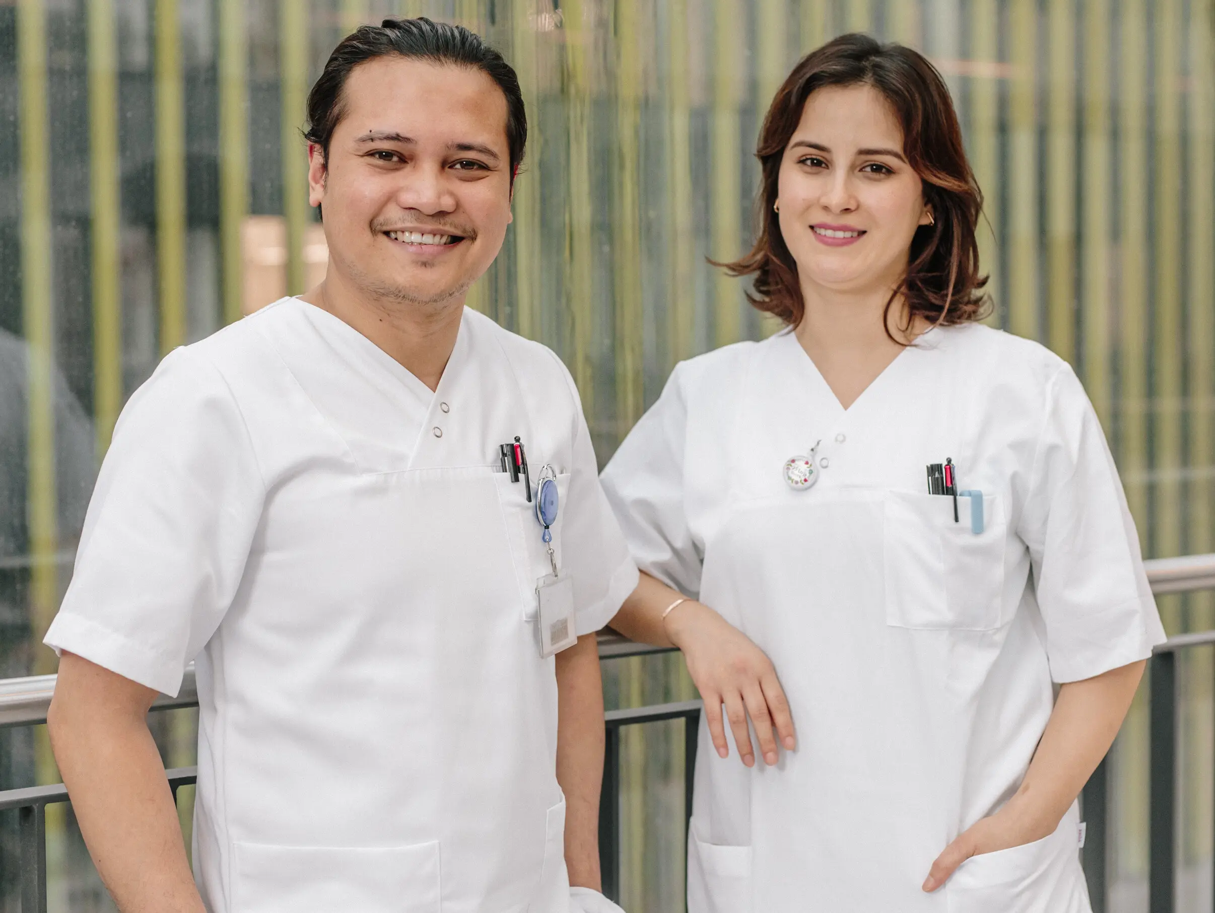 Dua profesional kesehatan yang tersenyum dengan jas putih berdiri di depan dinding kaca modern dengan garis-garis vertikal.