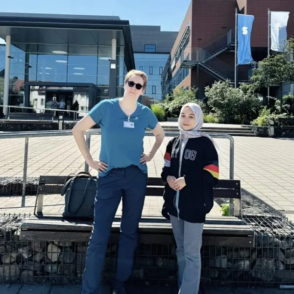 Dua orang berdiri di depan sebuah bangunan modern. Seorang wanita mengenakan kemeja biru dan kacamata hitam, sedangkan wanita lainnya mengenakan kaus hitam dengan garis-garis warna-warni. Keduanya tersenyum tipis.