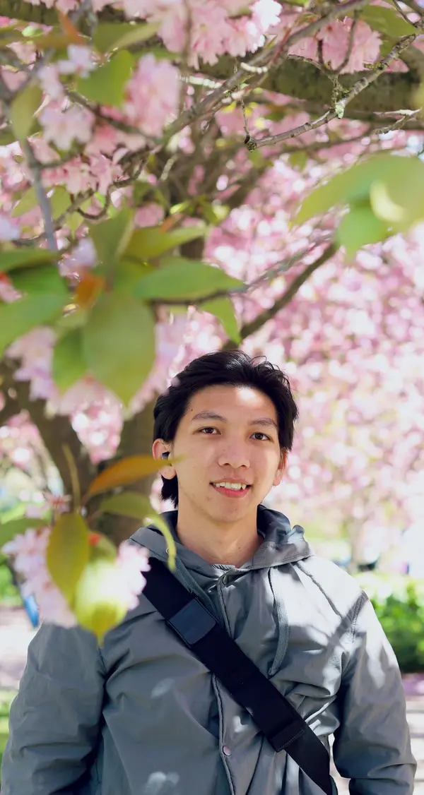 Seorang pemuda berdiri tersenyum di depan pohon sakura yang sedang mekar dengan bunga berwarna merah muda.