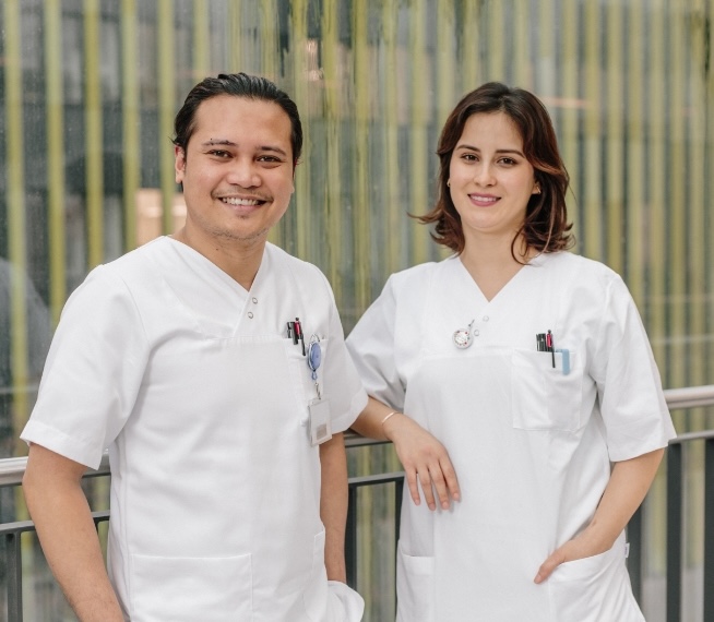 Dua orang profesional medis berbaju putih berdiri tersenyum di depan latar belakang yang modern.